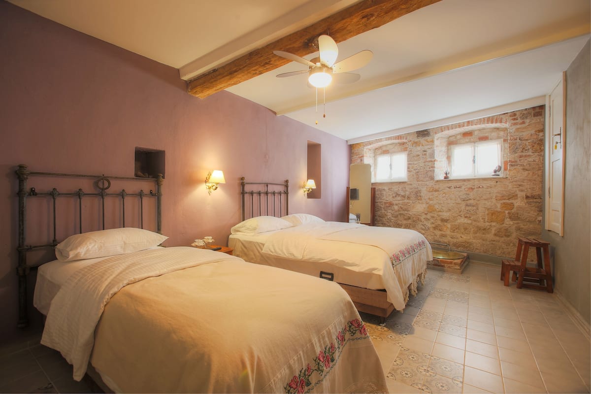A bedroom with two beds, a ceiling fan, stone walls, and a window with a view.