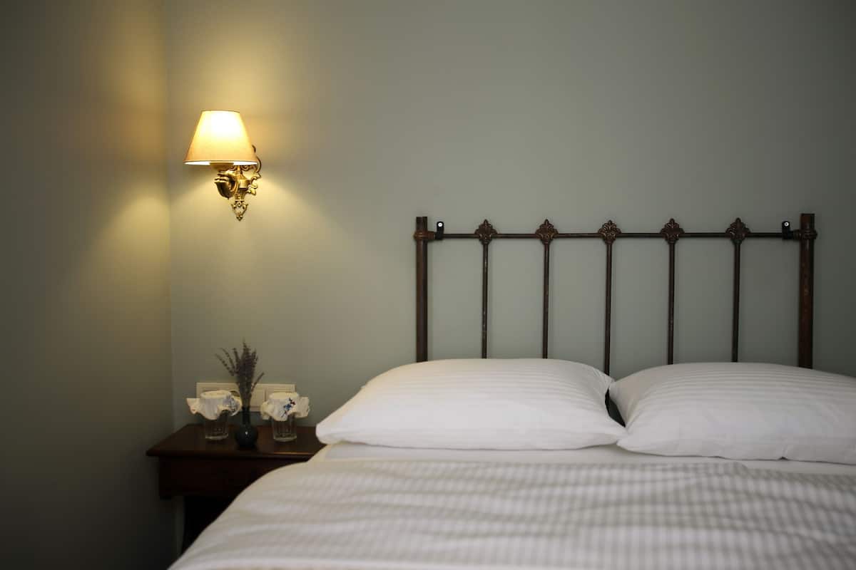 A neatly made bed with white linens, a wall-mounted lamp, and a small table with a vase and flowers.