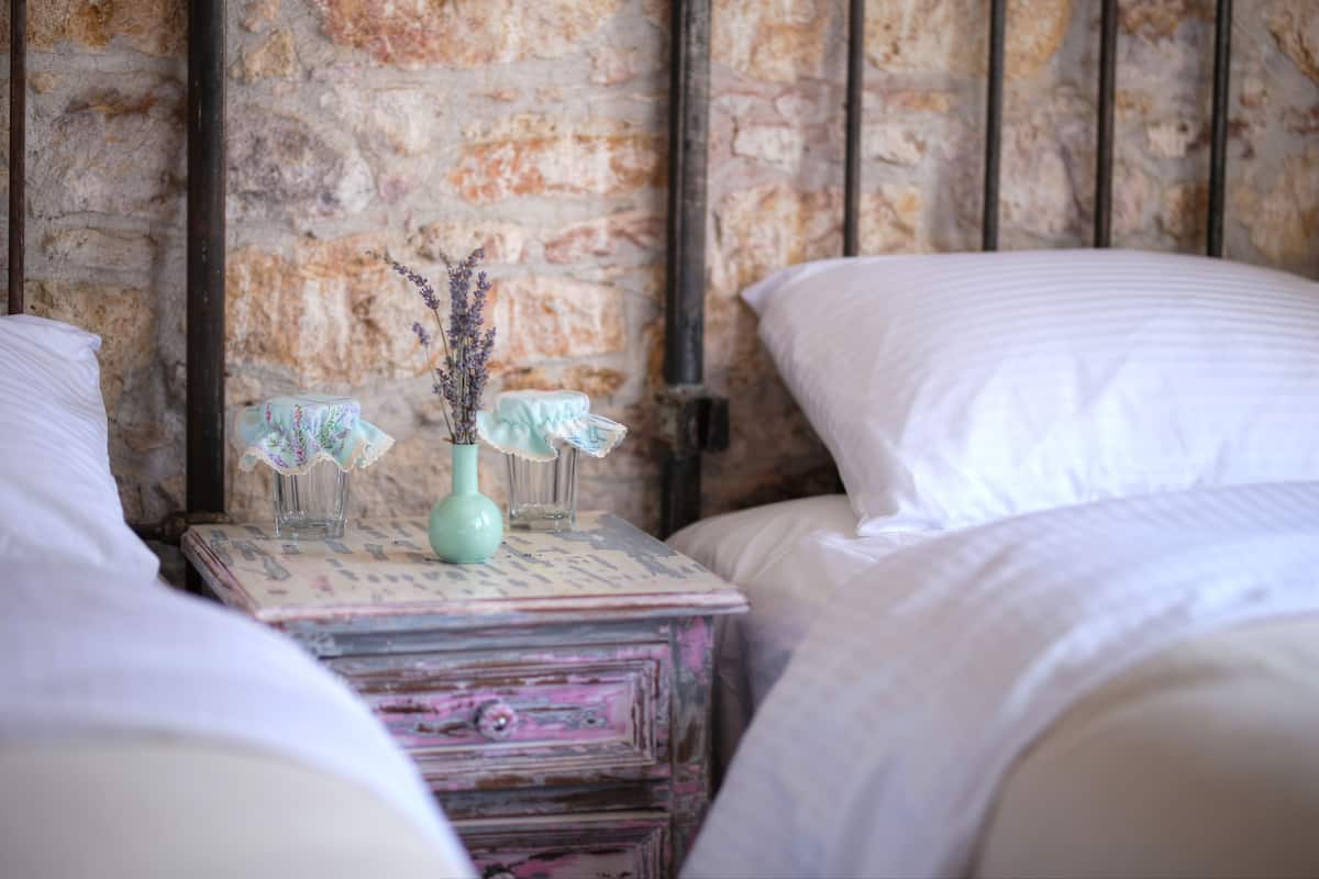 A bedroom with a rustic brick wall, a vintage dresser with a vase, and white bedding.