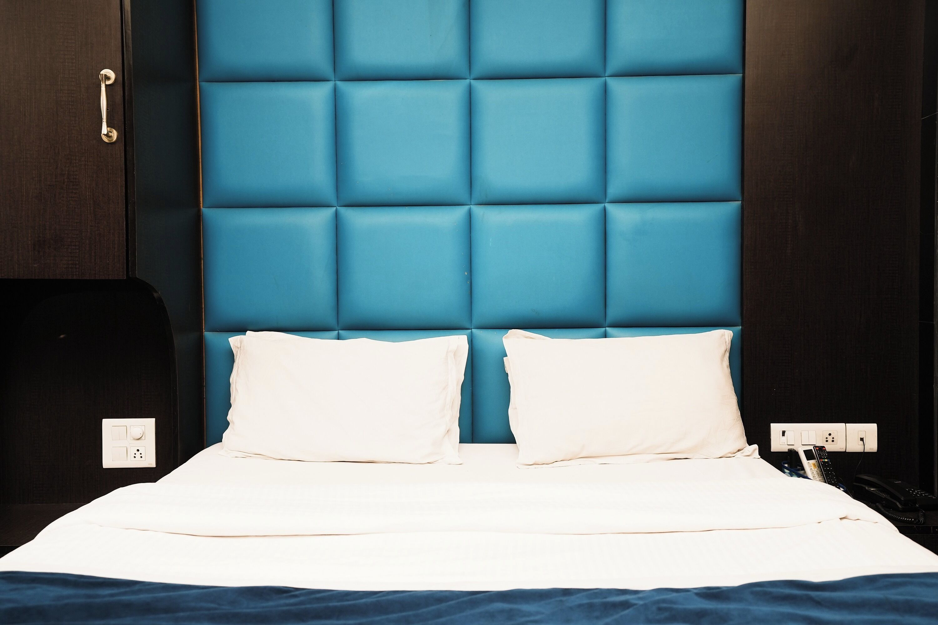 Ein Bett mit blauem Kopfteil, weißer Bettwäsche und einem Nachttisch mit Telefon und Ladegerät.