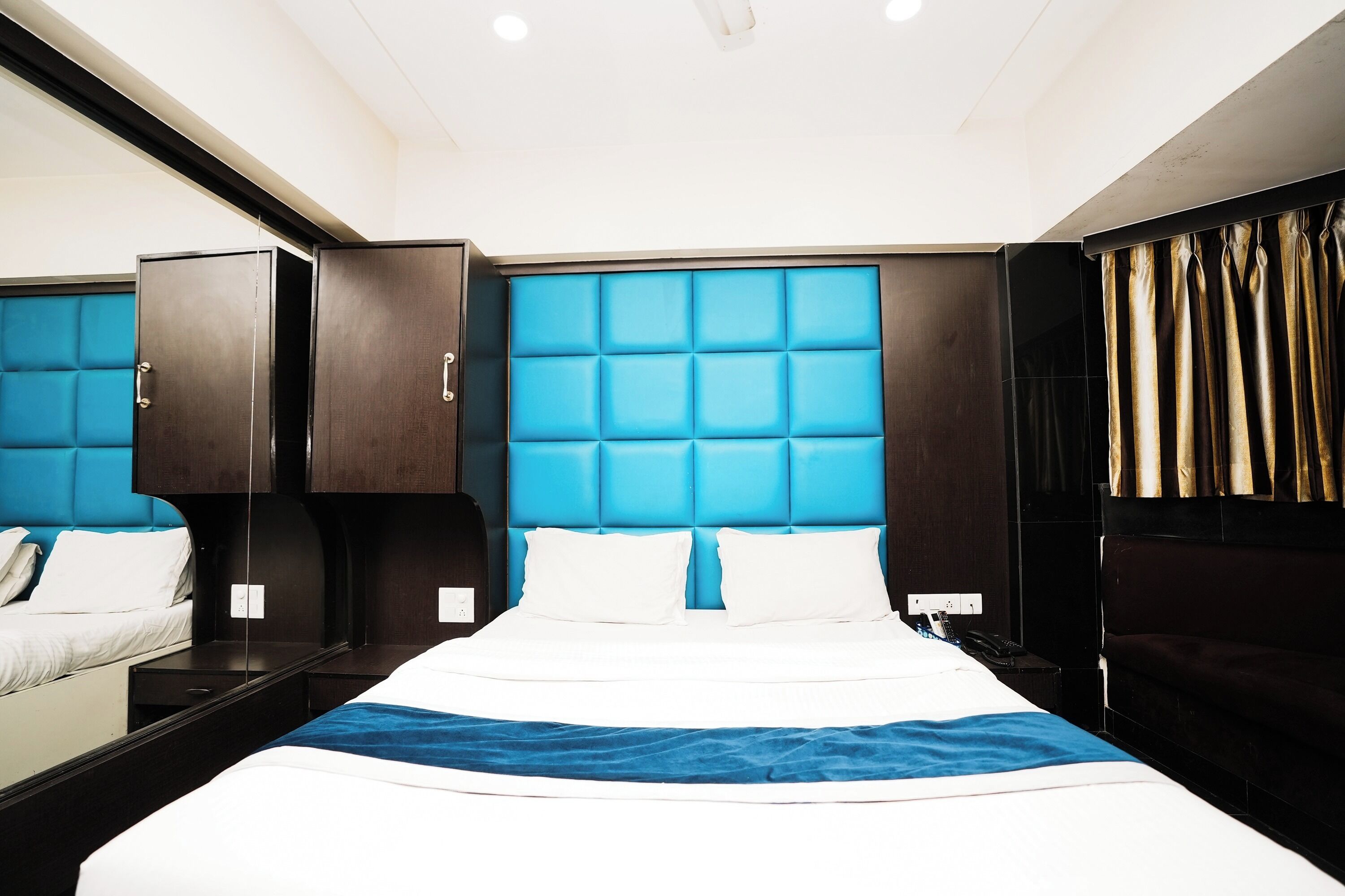 A hotel room with a large bed, a headboard with a blue padded design, a wardrobe, and a nightstand with a lamp.