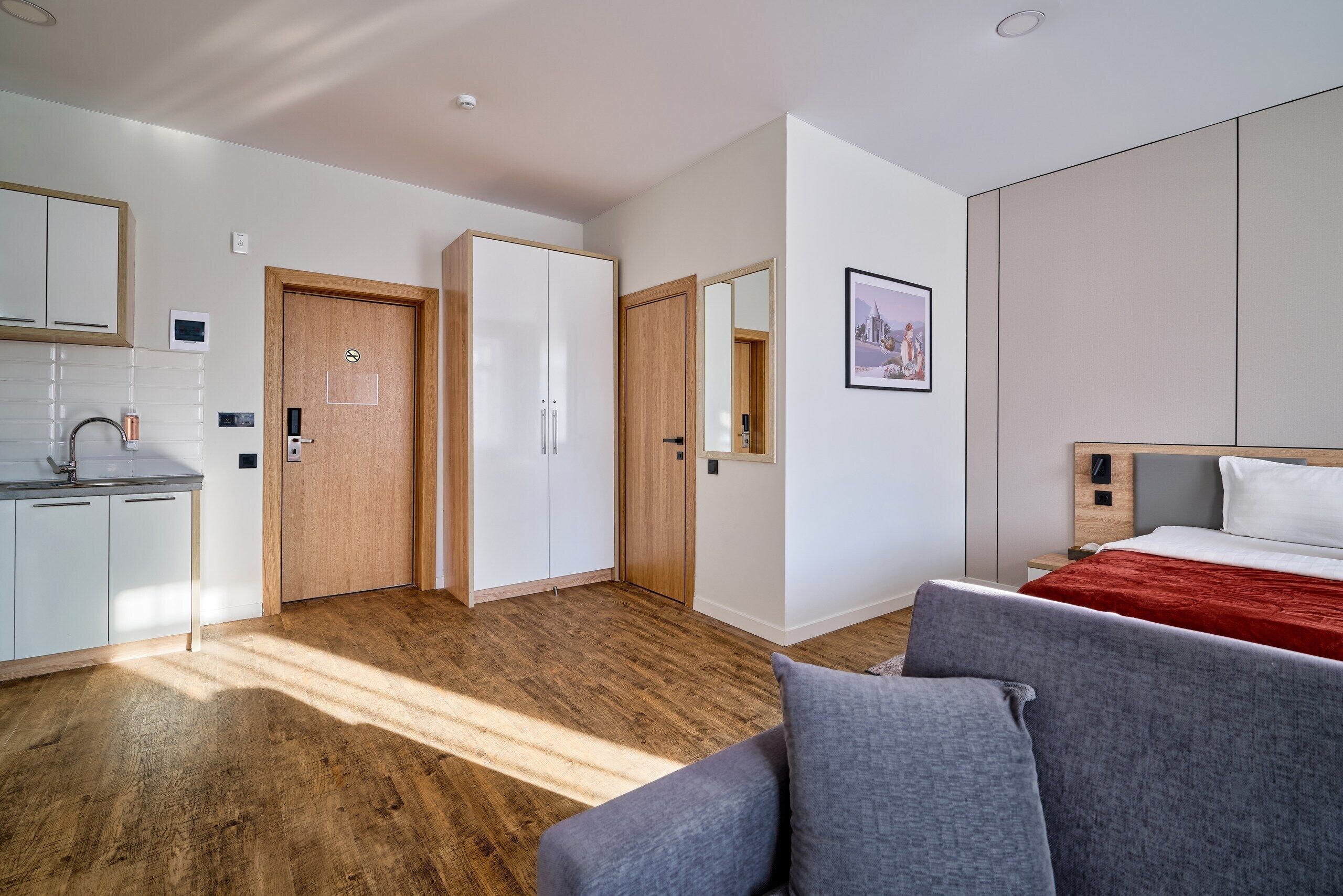 Una camera d'albergo moderna con pavimento in legno, un letto con coperta rossa, un divano grigio, un armadio in legno, un mobiletto bianco e uno specchio a parete.