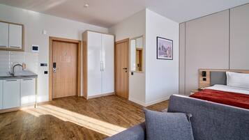 Una camera d'albergo moderna con pavimento in legno, un letto con coperta rossa, un divano grigio, un armadio in legno, un mobiletto bianco e uno specchio a parete.