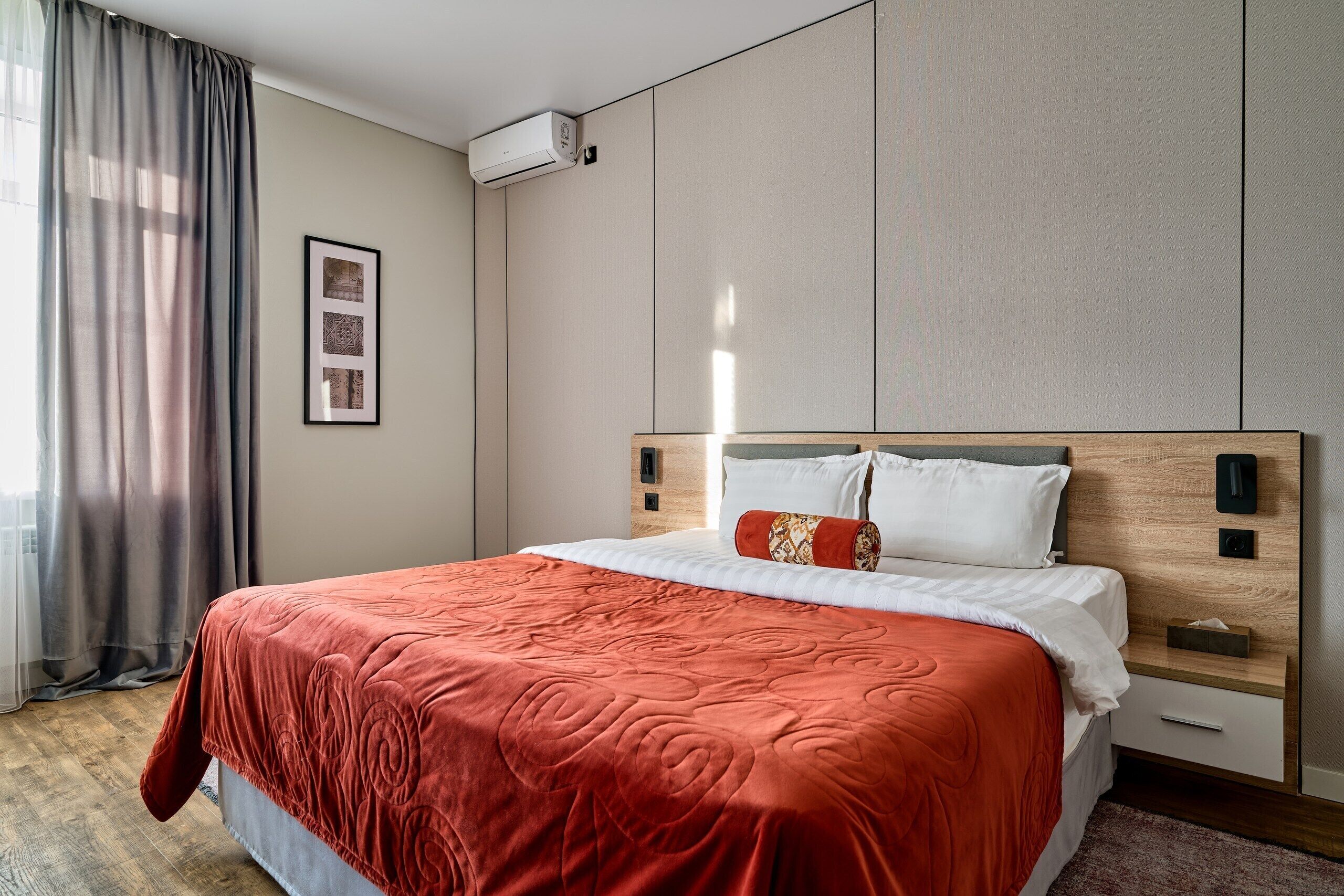 Een net opgemaakt bed met een rode deken, een nachtkastje met een lamp, en een wandairconditioner.