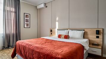 Een net opgemaakt bed met een rode deken, een nachtkastje met een lamp, en een wandairconditioner.