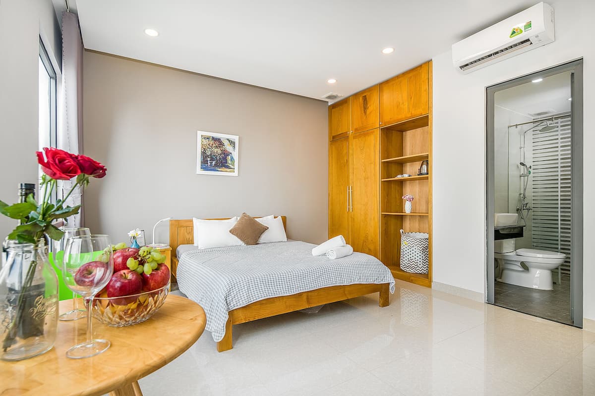 A modern bedroom with a wooden bed, a bedside table with a fruit bowl, a vase with a red rose, and a wall-mounted air conditioner.
