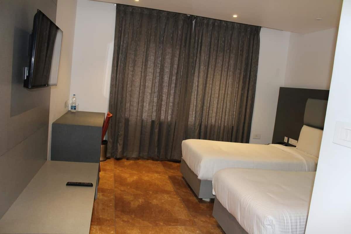 A hotel room with two beds, a TV mounted on the wall, a small table with a bottle, and a curtain.