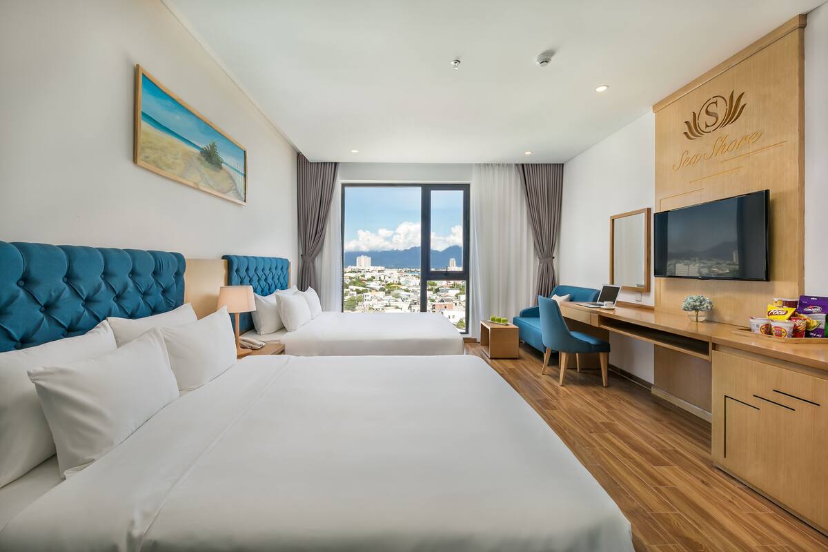 A hotel room with two beds, a desk, a TV, and a large window with a city view.