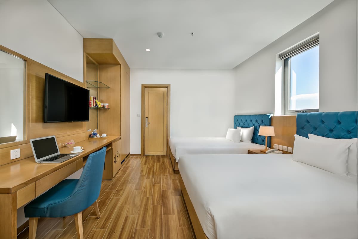 A hotel room with a wooden desk, a laptop, a blue chair, two beds with white bedding, a blue headboard, a window with blinds, and a door.