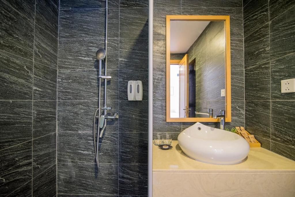 Een badkamer met een modern design, met een douche met handdouche en een muurbevestigde kraan, een witte wastafel met een beige aanrechtblad en een spiegel met houten lijst.
