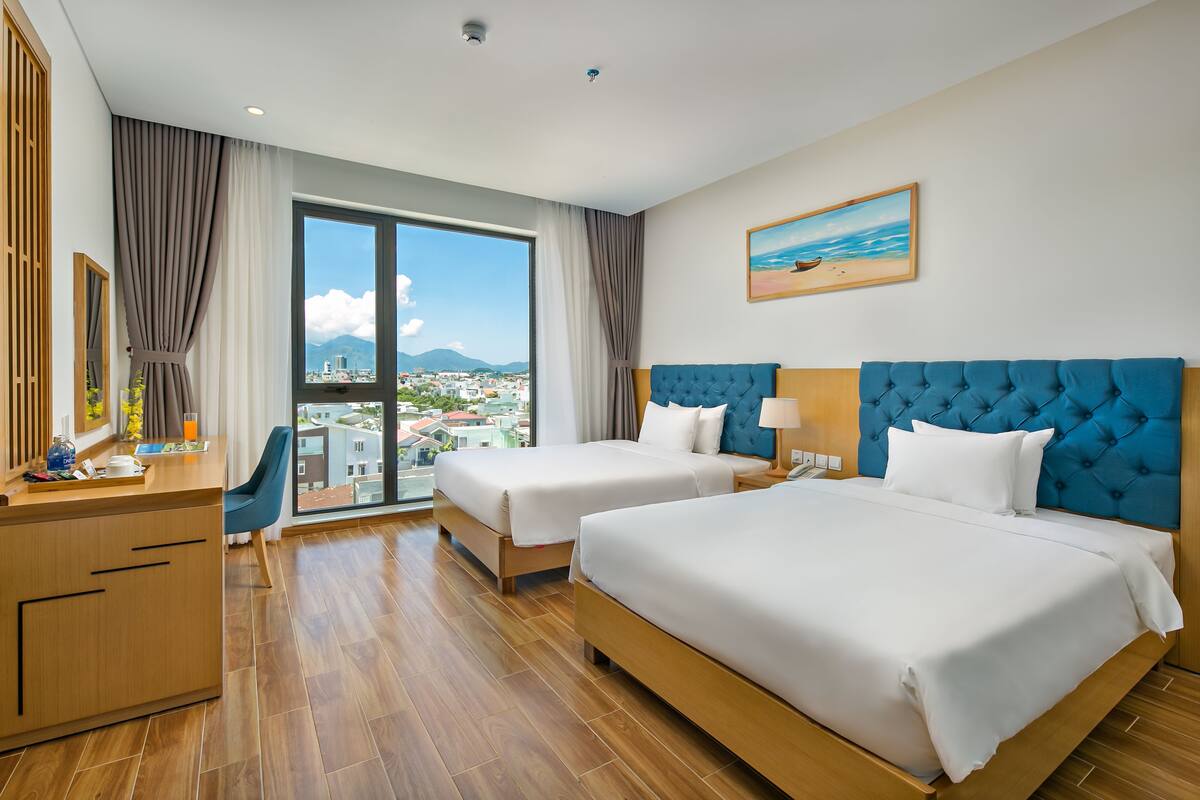 A hotel room with two beds, a desk, and a large window with a city view.