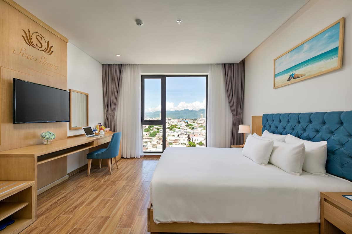 A hotel room with a large bed, a desk with a laptop, a flat-screen TV, and a city view through the window.