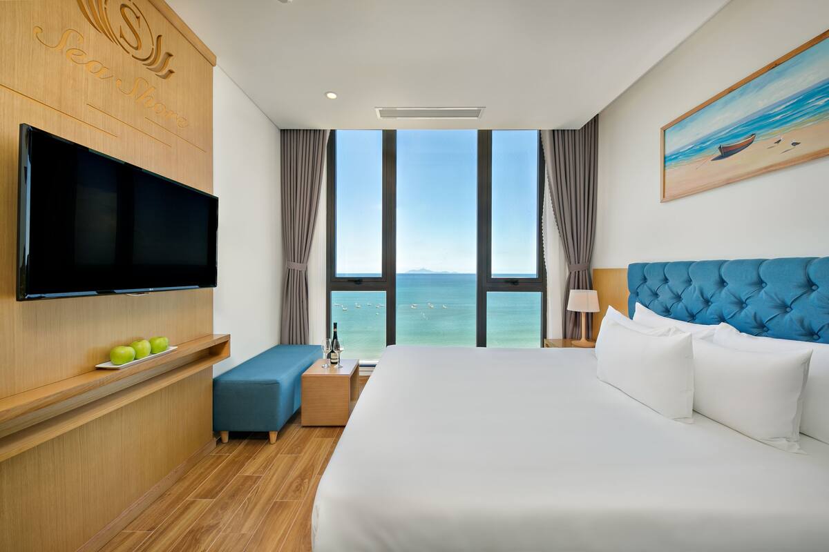A hotel room with a large bed, a flat-screen TV, a wooden desk, and a view of the sea.