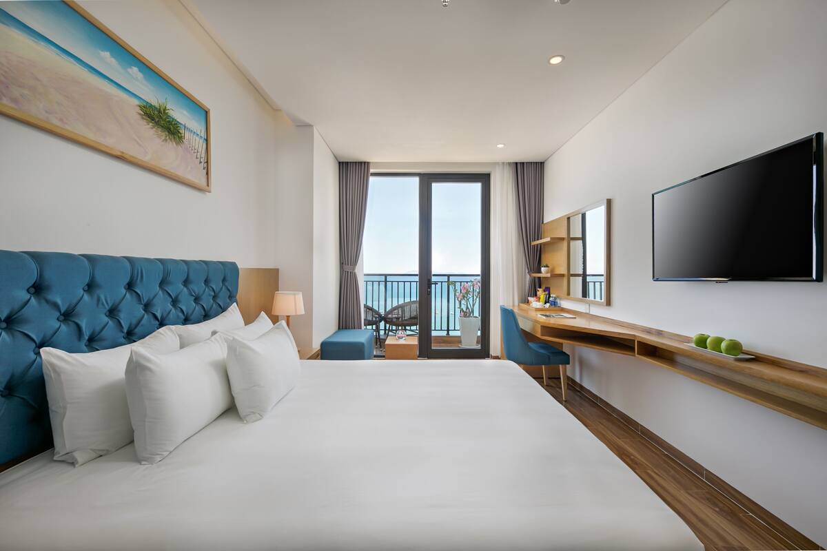 A hotel room with a large bed, a desk, a TV, and a balcony with a view.