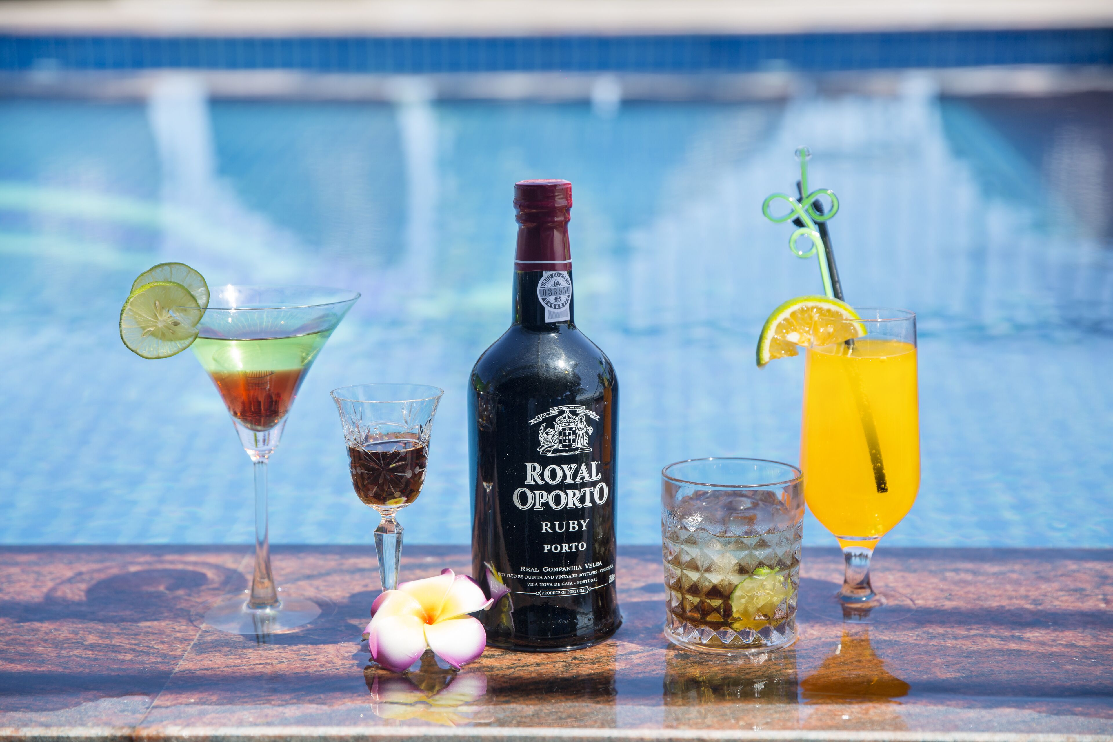 A bottle of Royal Oporto Ruby Porto, two glasses of cocktails, and a glass of orange juice on a table by a pool.