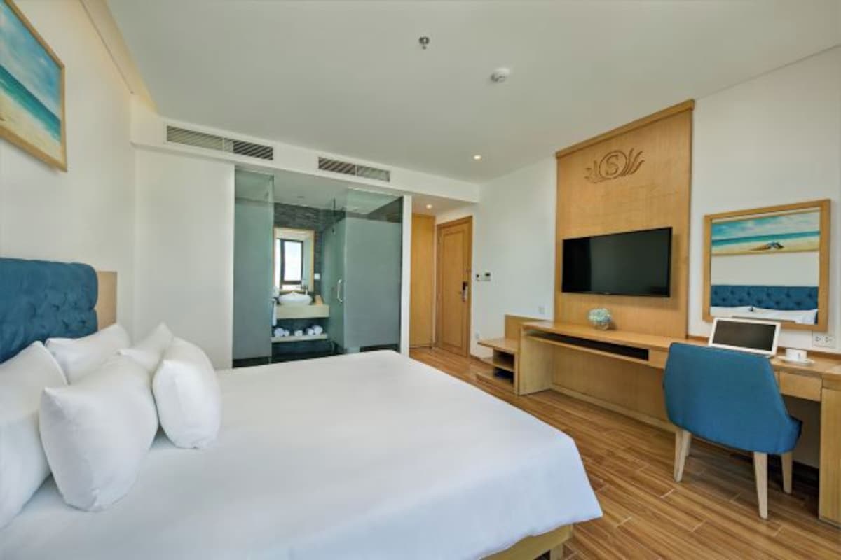 A modern hotel room with a large bed, a desk with a computer, a TV, and a view of the bathroom.