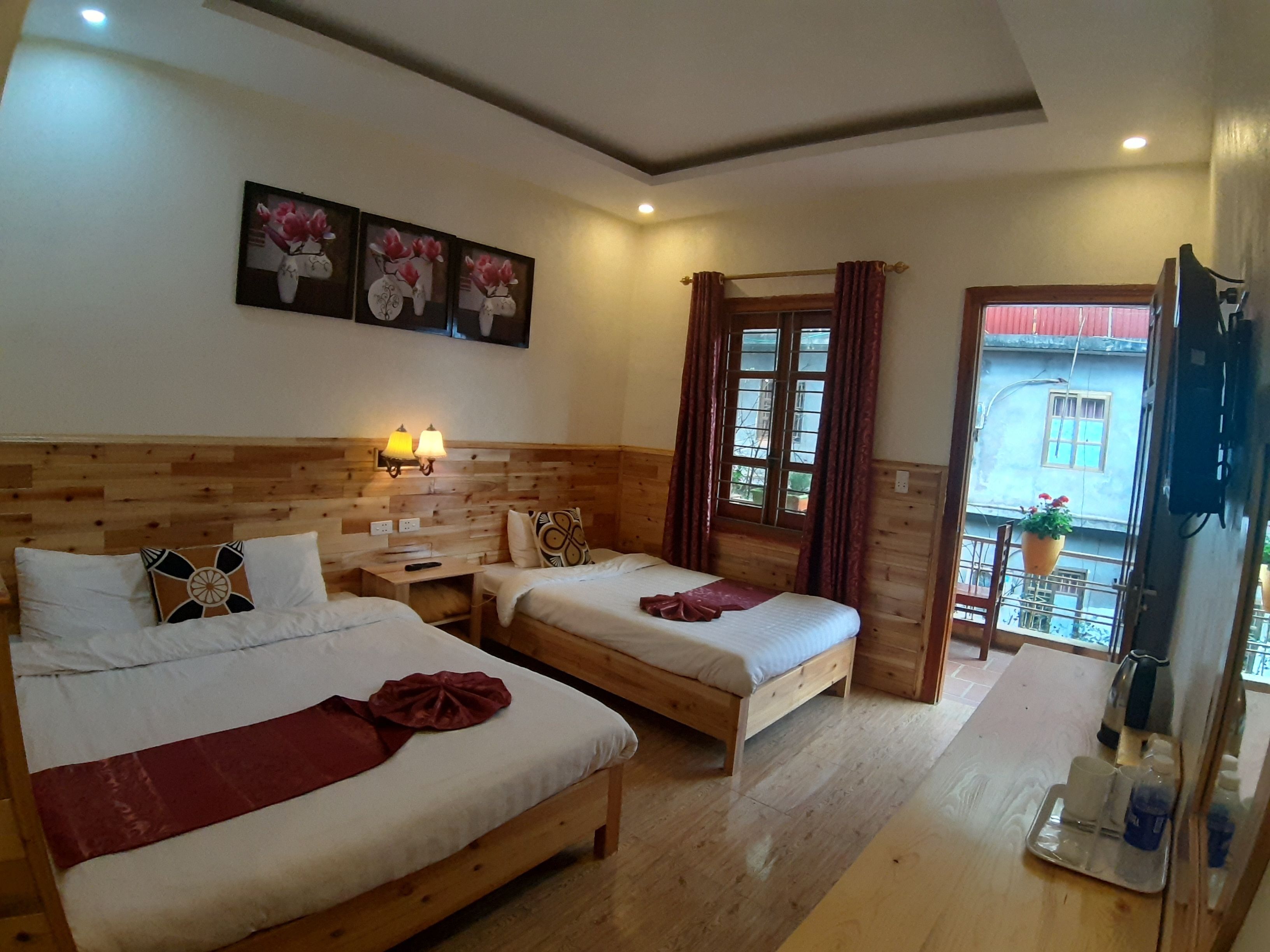 A hotel room with two beds, wooden headboard, wall-mounted lamps, a window with curtains, and a balcony with a hanging planter.