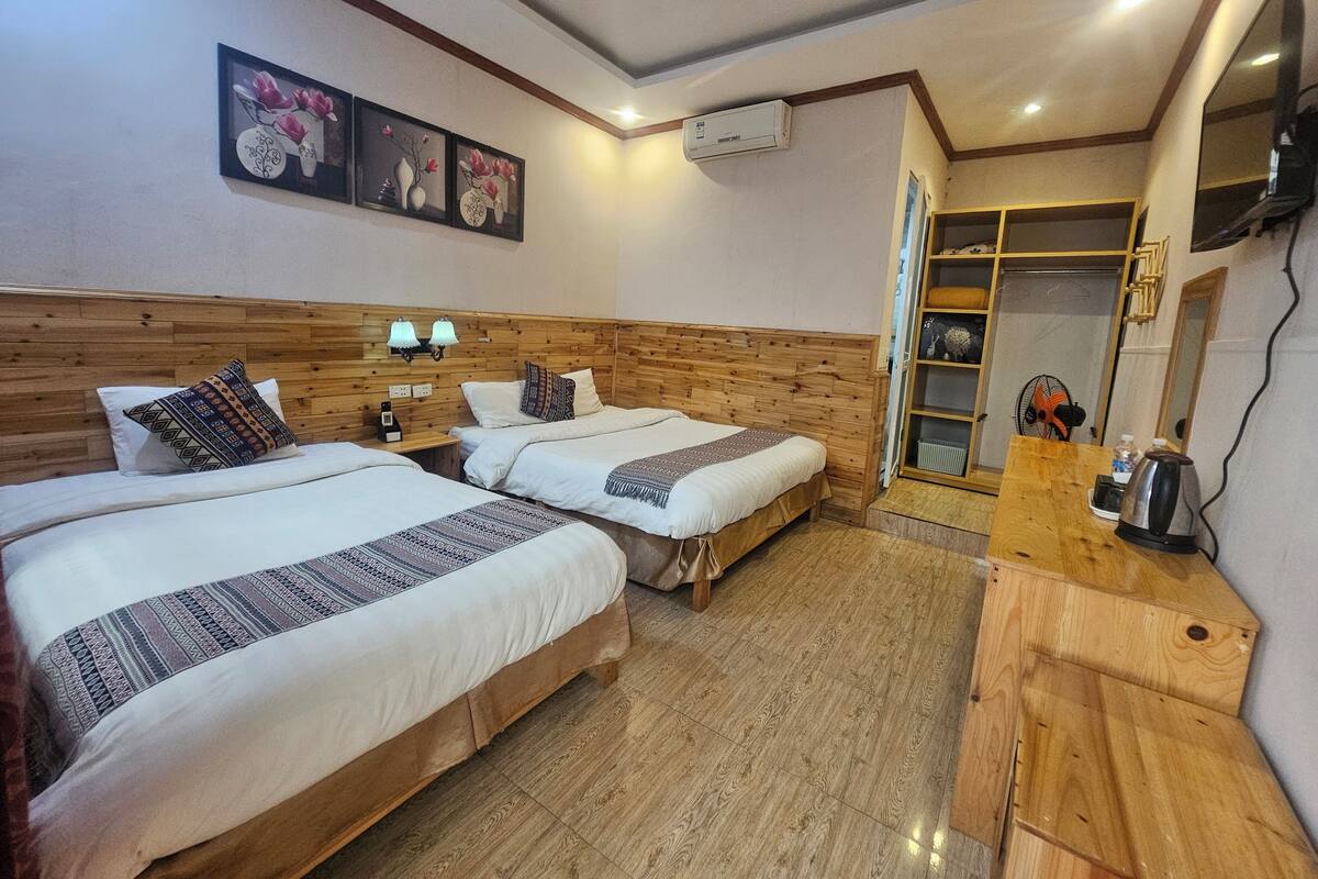 A hotel room with two beds, wooden headboard, wall-mounted air conditioning unit, and a wooden desk with a kettle.