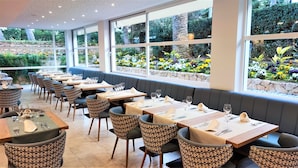 Ein Restaurant mit mehreren Tischen und Sitzecken, großen Fenstern mit Gartenblick und einer Steinsäule.