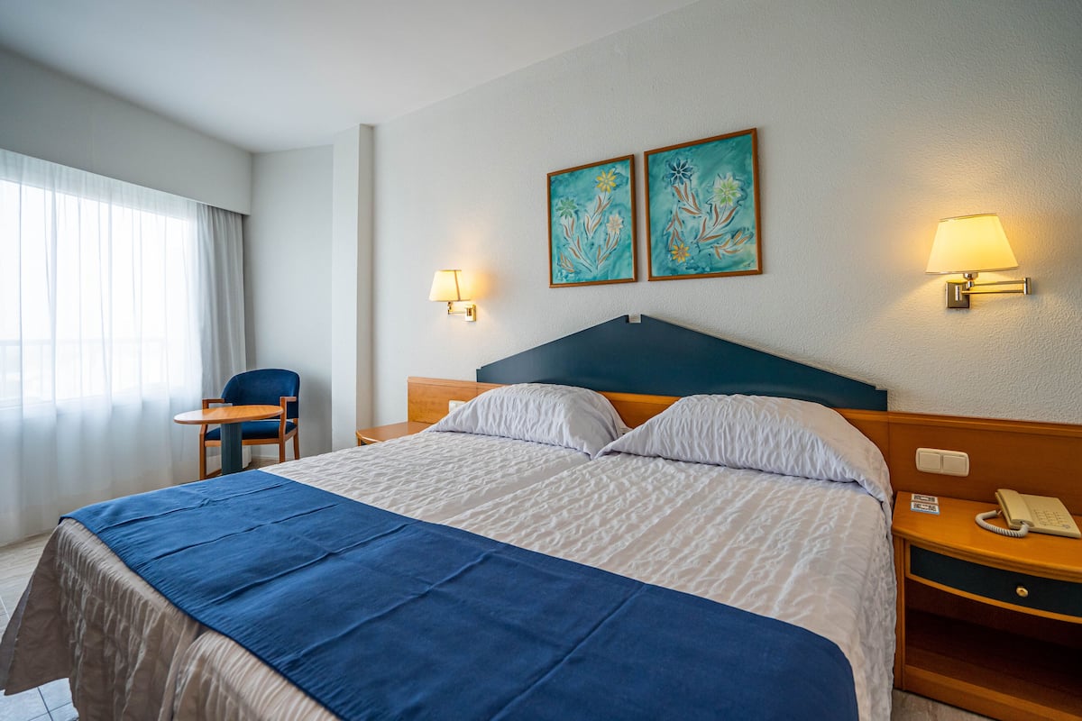 Une chambre d’hôtel comprenant un lit, une chaise, une table de chevet avec un téléphone et deux lampes fixées au mur.