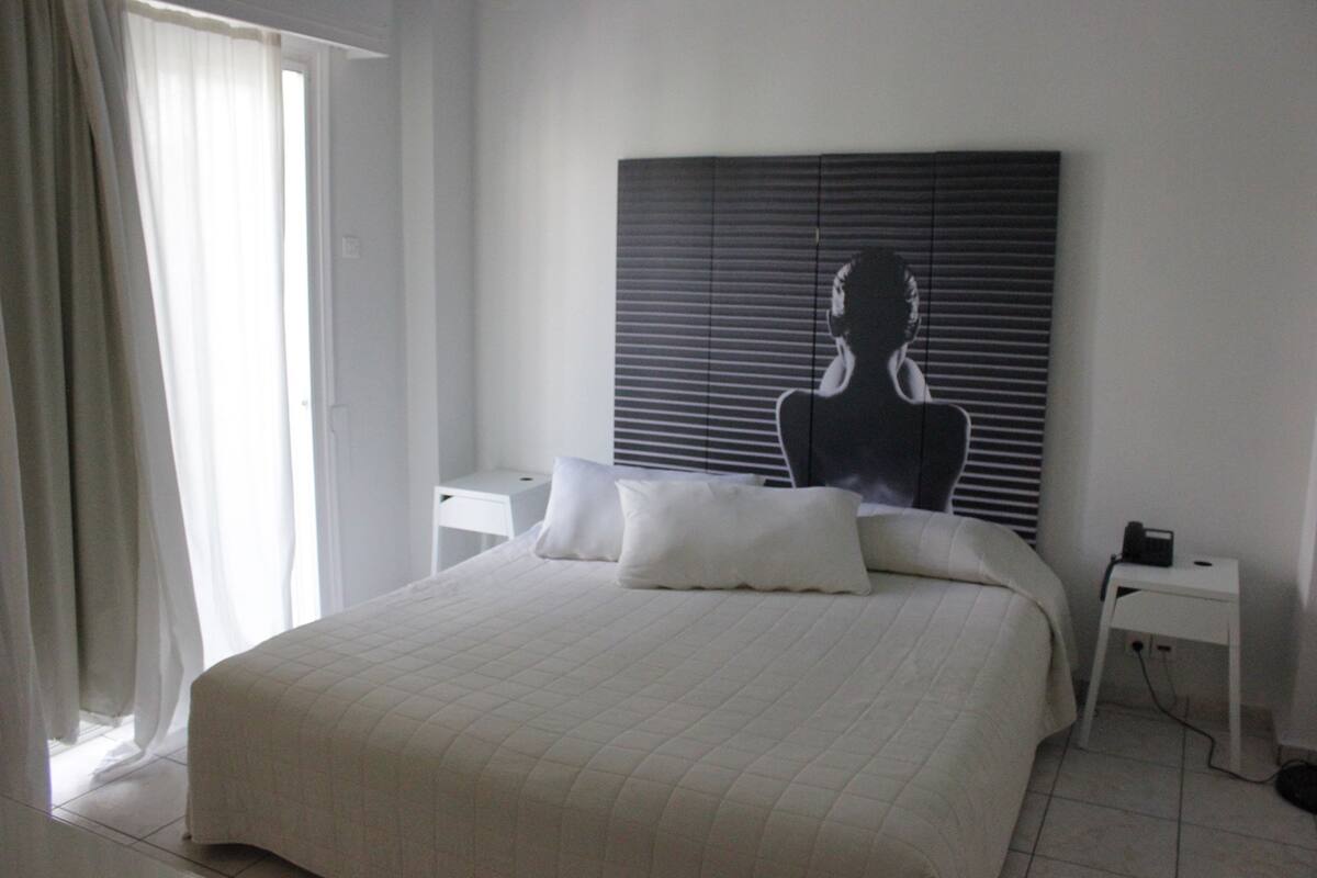 A bedroom with a large bed, white bedding, a nightstand, a lamp, and a window with curtains.