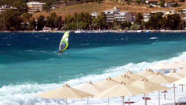 Een kustgezicht met een windsurfplank, strandparasols en gebouwen op de achtergrond.