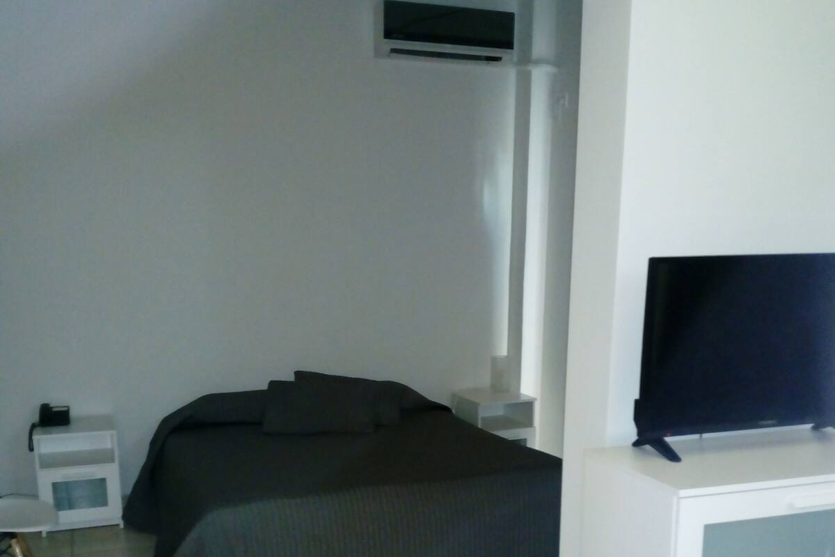 A bedroom with a bed, a TV on a stand, a bedside table, and a wall-mounted air conditioning unit.