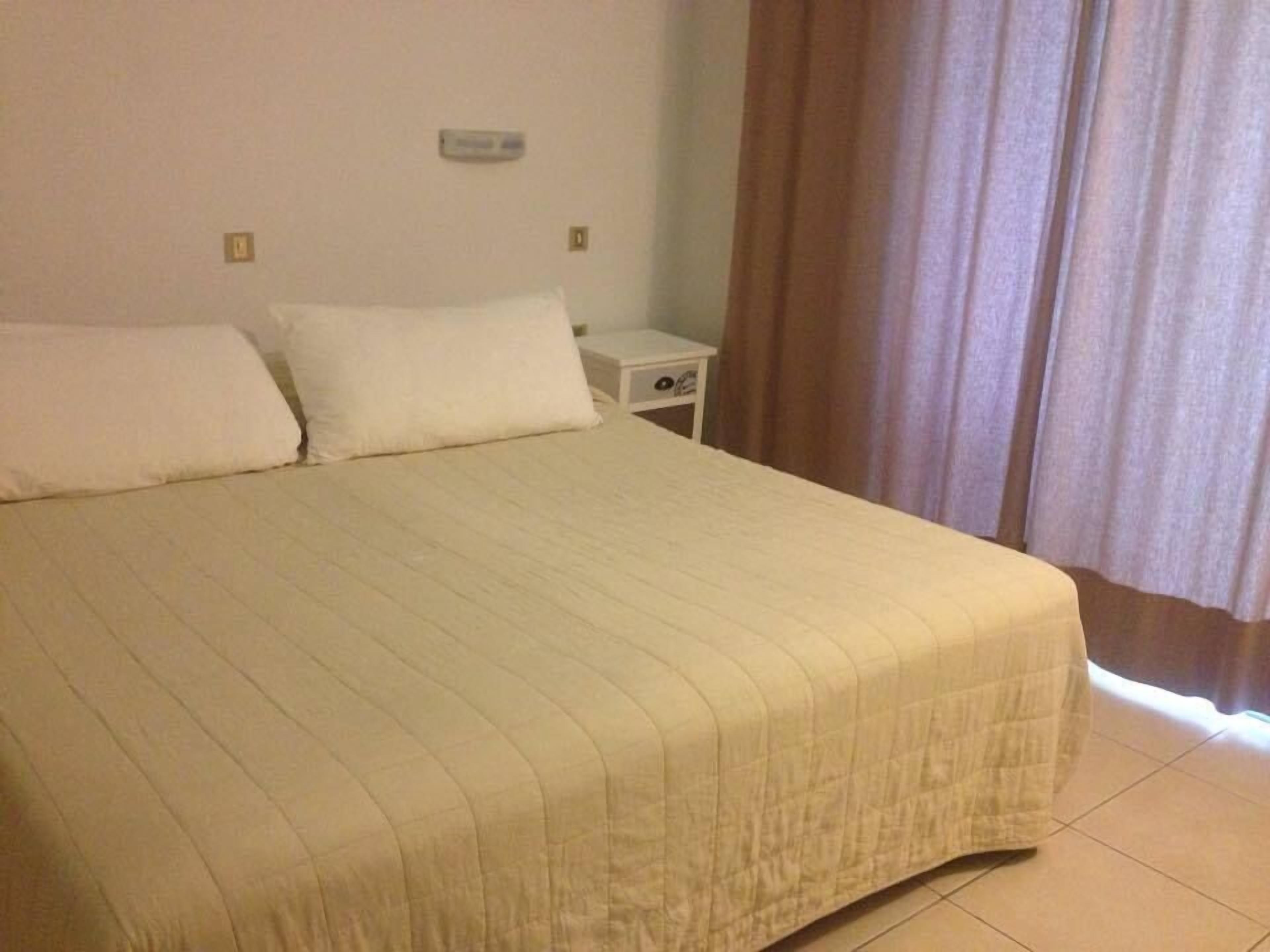 Un lit bien fait, recouvert d’une courtepointe matelassée, une table de chevet avec une lampe et une fenêtre avec des rideaux.