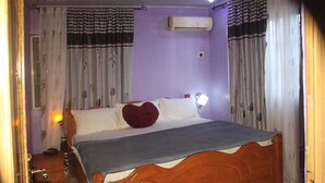 A bedroom with a wooden bed, white bedding, a red heart pillow, and an air conditioning unit on the wall. - Hotel Princebella (Lagos)