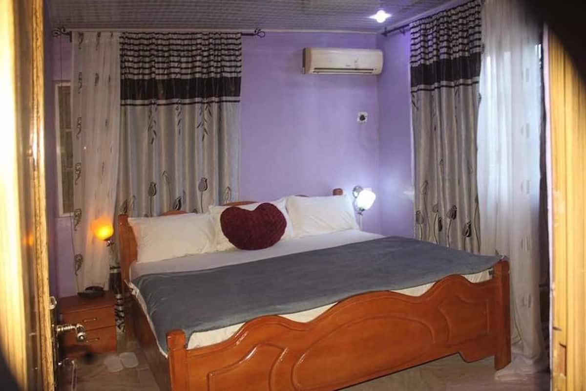 Un dormitorio con cama de madera, ropa de cama blanca, un cojín con forma de corazón rojo y un equipo de aire acondicionado en la pared.