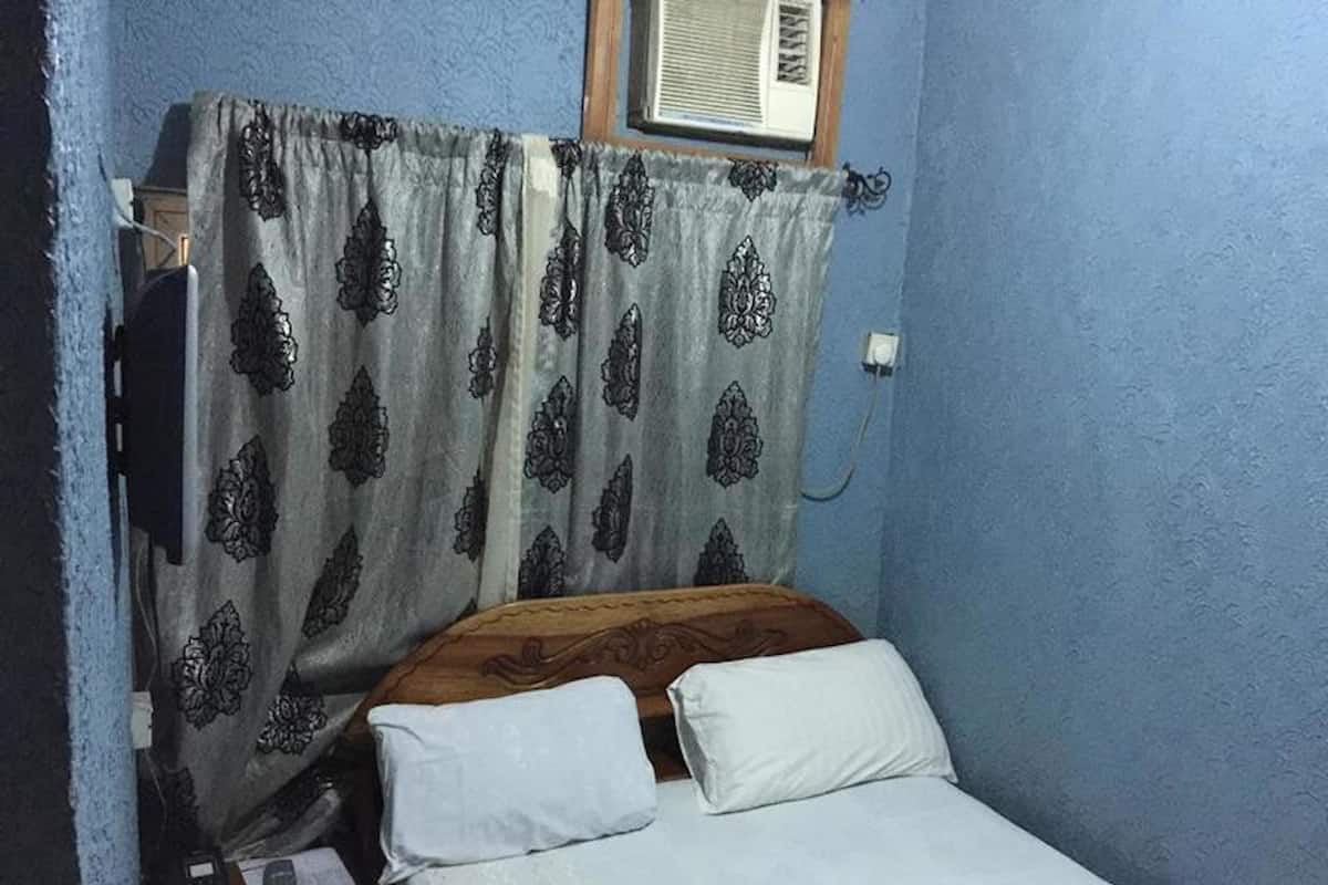 Una habitación pequeña con cama individual, cabecera de madera, mesita de noche con lámpara y ventana con cortina.