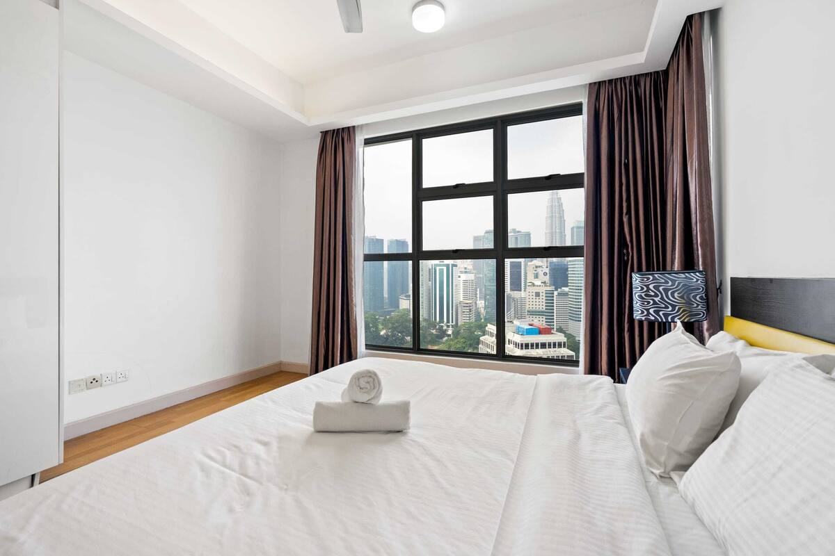 A hotel room with a large window, a bed with white linens, a headboard, and a view of the cityscape.