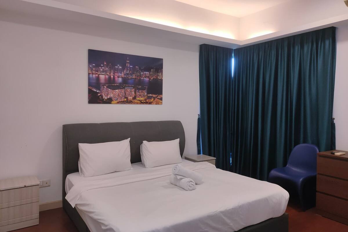 A neatly made bed with white linens, a cityscape painting above it, a dark blue curtain, and a blue chair.