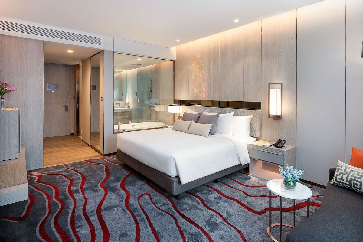 A modern hotel room with a large bed, a bedside table with a lamp, a small round table with a vase, and a bathroom visible through a glass door.