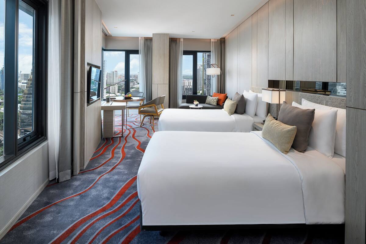 A hotel room with two beds, a desk, a chair, a TV, and a view of the city.