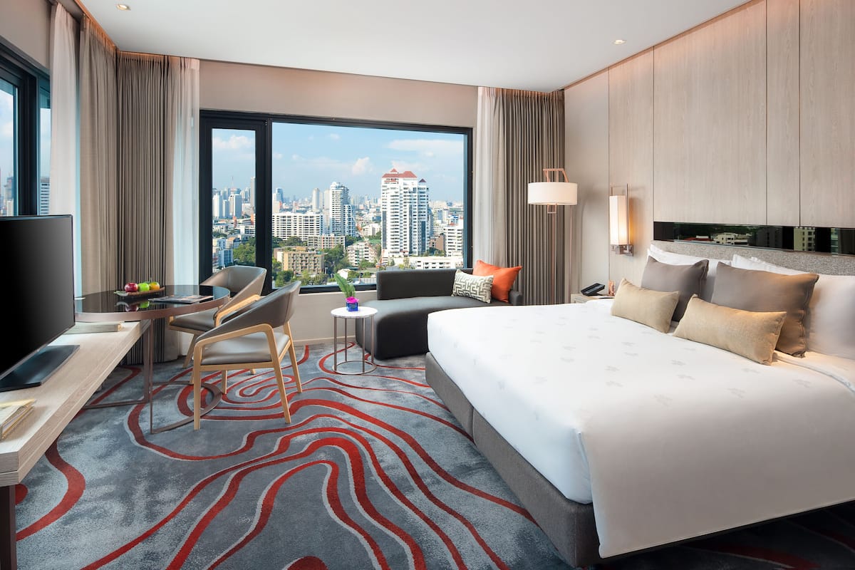 A modern hotel room with a large bed, a desk with a flat-screen TV, a chair, a small side table, and a view of the city skyline.