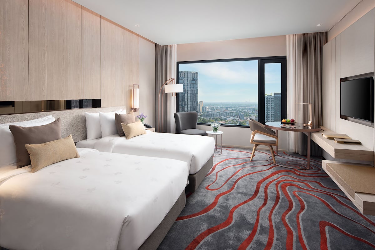 A hotel room with two beds, a desk, a chair, a TV, and a large window with a city view.