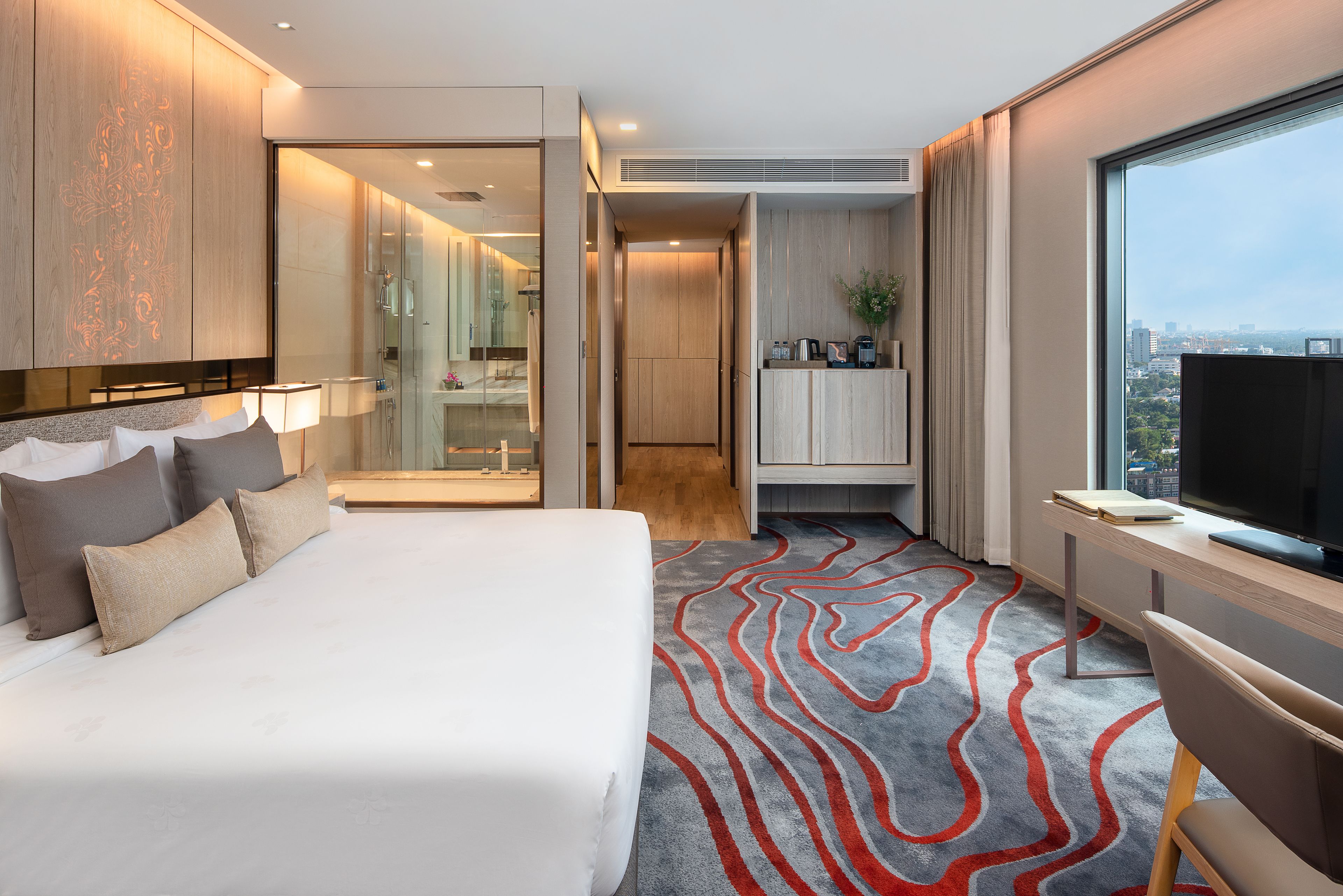 A modern hotel room with a large bed, a desk with a TV, a view of the city, and a shower area.