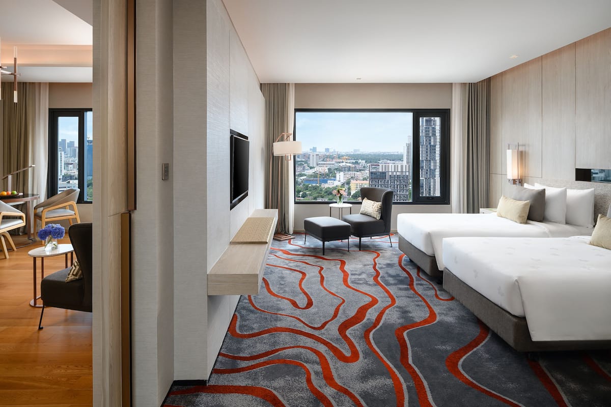 A modern hotel room with a large window, a flat-screen TV, a sofa, a bed, and a small table with a vase of flowers.