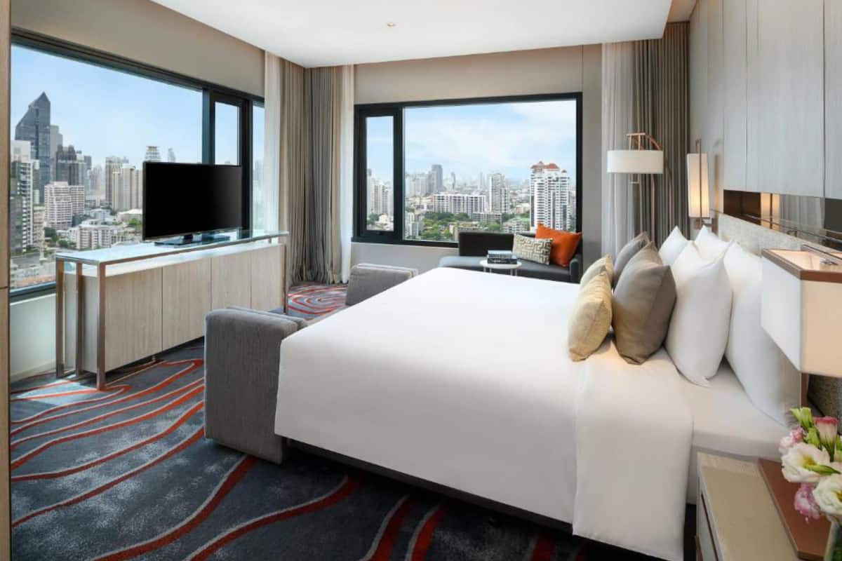 A hotel room with a large bed, a desk with a TV, and a view of the cityscape through large windows.