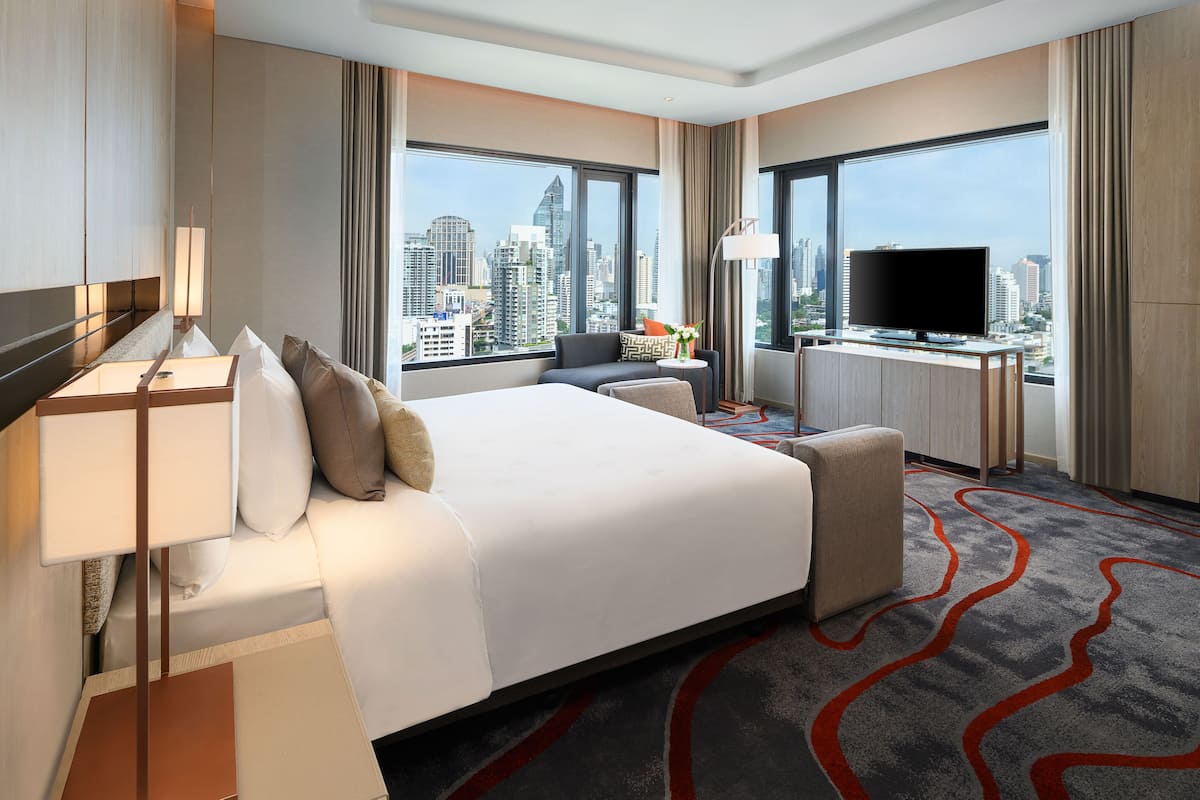 A hotel room with a large bed, a desk with a flat-screen TV, a sofa, and a view of the city skyline.