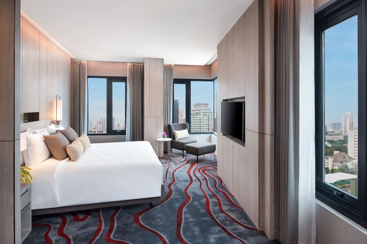 A modern hotel room with a large bed, a TV, a small table with a chair, and a view of the city through large windows.