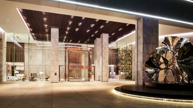 Um hall de hotel moderno, com entrada ampla, uma coluna central e uma grande escultura metálica.