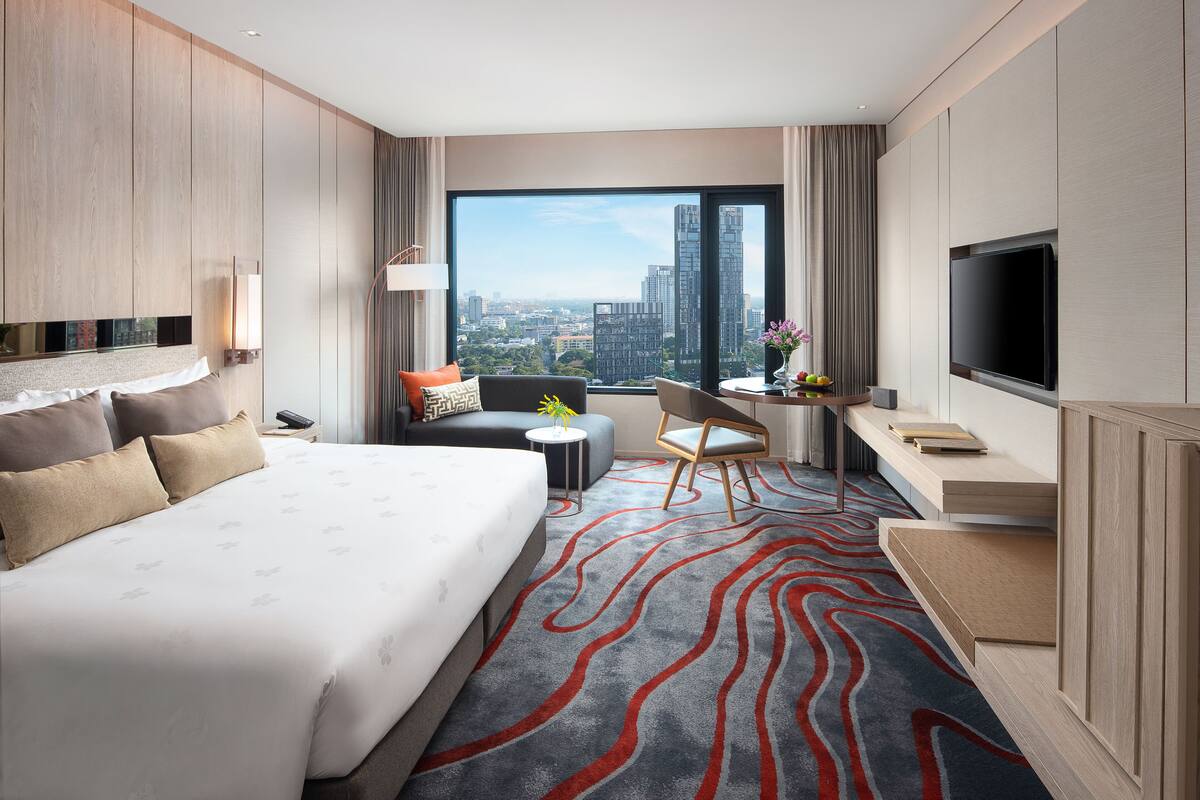 A modern hotel room with a large bed, a desk with a chair, a TV, and a view of the city.