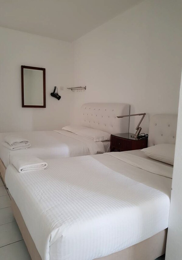 A hotel room with two beds, a bedside table with a lamp, a wall-mounted mirror, and a towel rack.