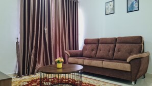 A living room with a brown sofa, a round coffee table with a yellow flower vase, a patterned rug, and a large window with curtains. - Anjung Apartment 3BR 1 (Banting)