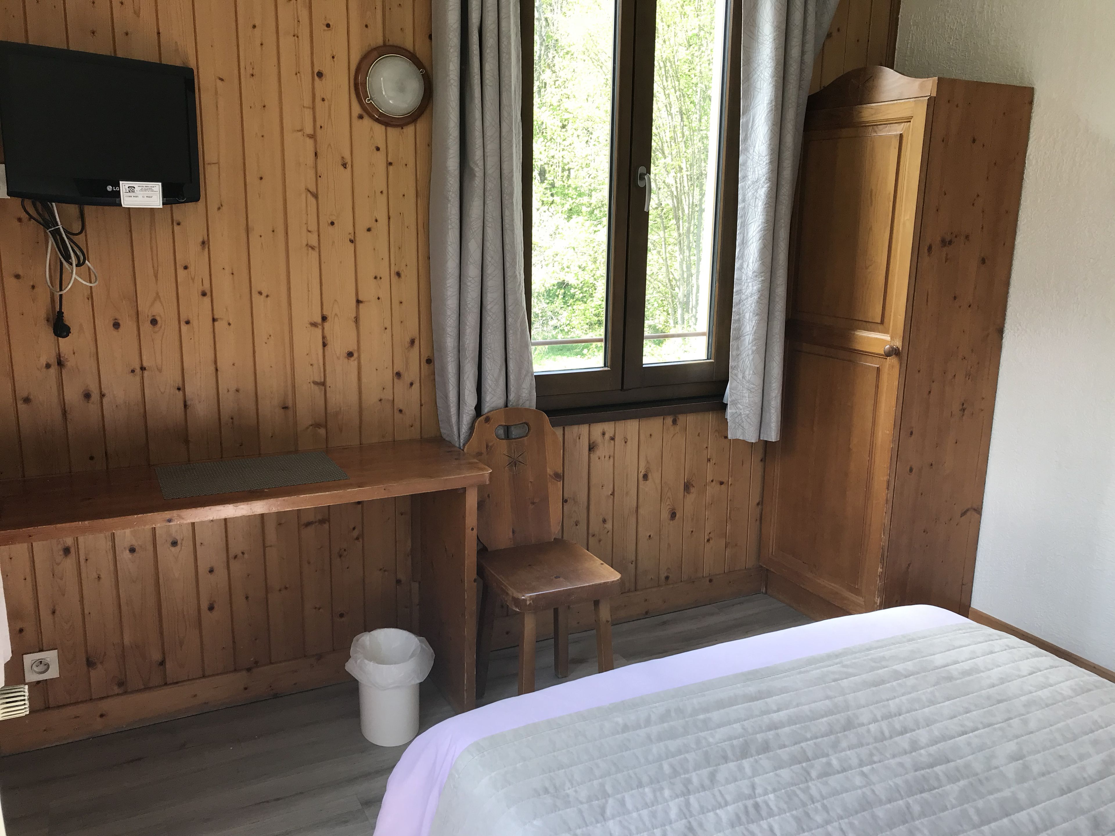 Una habitación de madera con cama, escritorio, silla, televisor y ventana con cortinas.