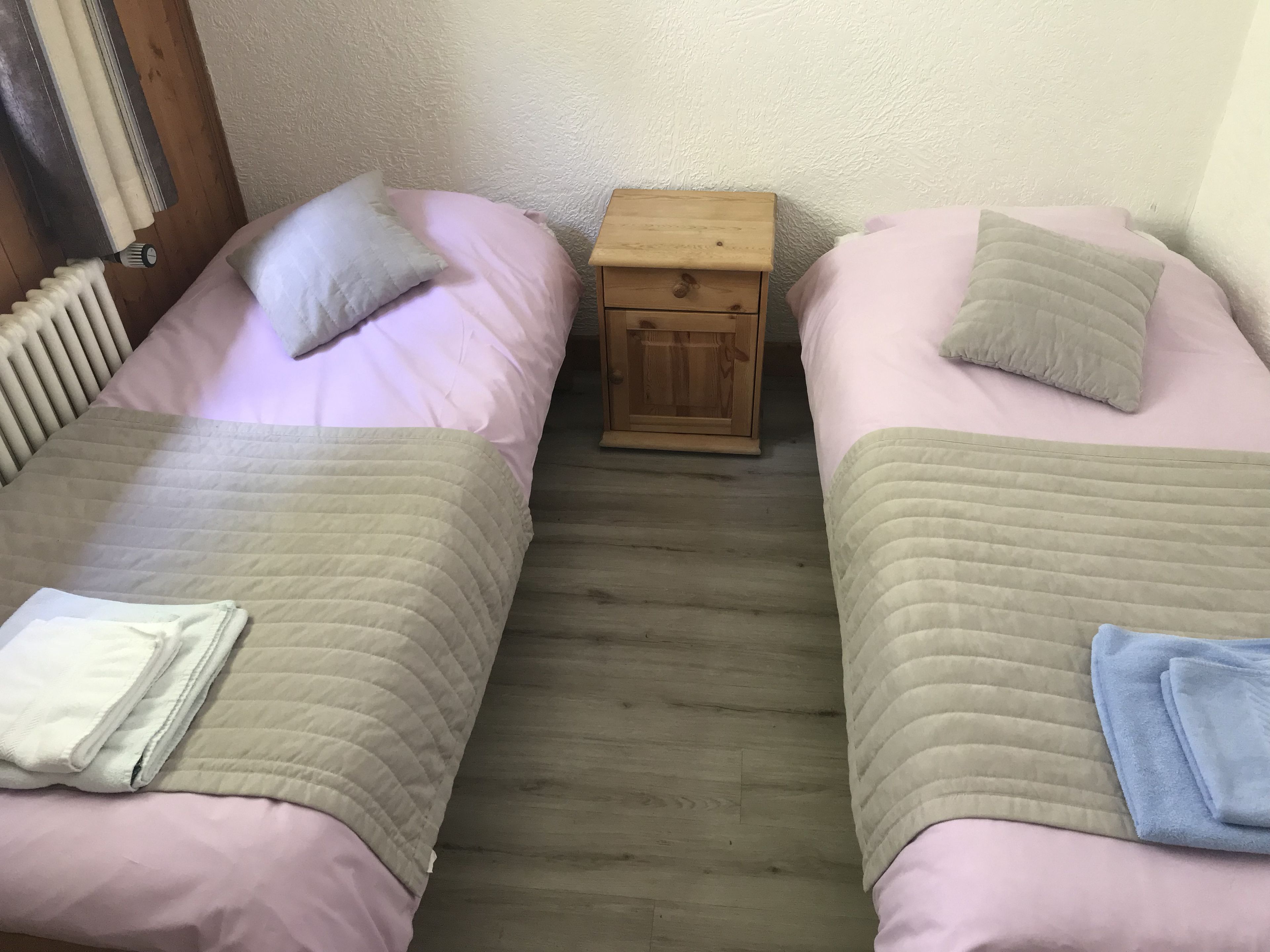 Ein kleiner, gemütlicher Raum mit zwei Betten, jeweils mit Kissen und einem gefalteten Handtuch. Dazwischen steht ein Nachttisch aus Holz.