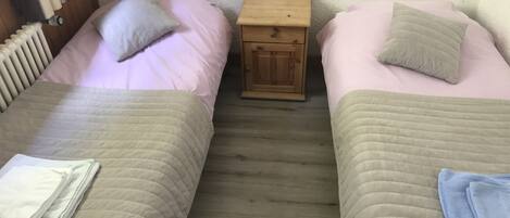 Ein kleiner, gemütlicher Raum mit zwei Betten, jeweils mit Kissen und einem gefalteten Handtuch. Dazwischen steht ein Nachttisch aus Holz.
