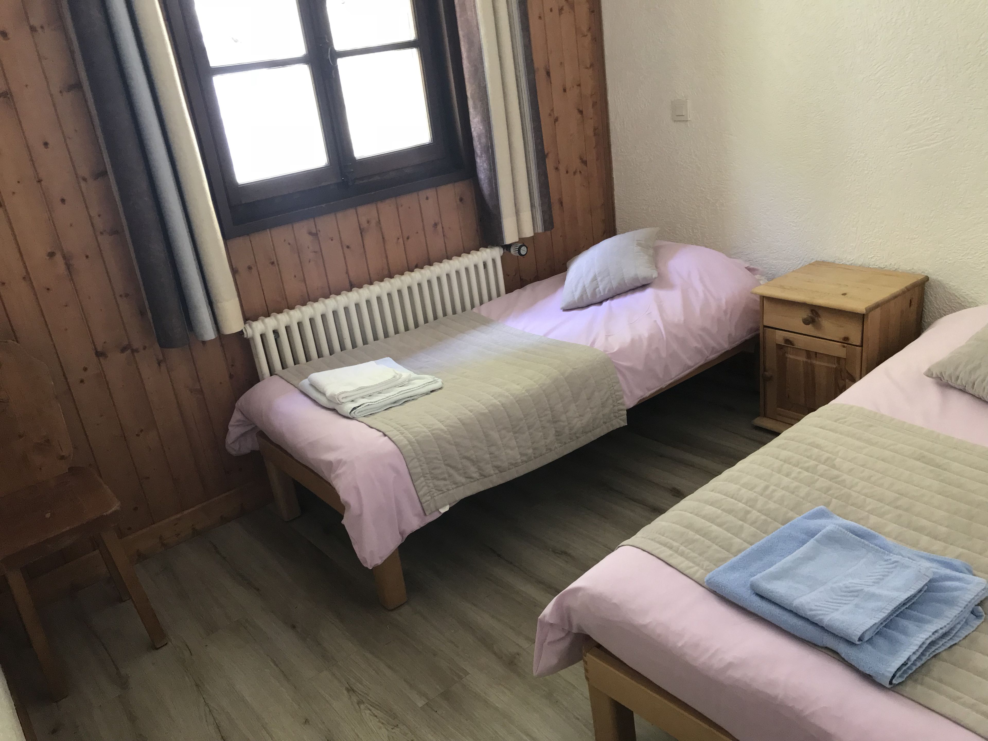 A small, cozy room with two single beds, a wooden floor, a radiator, and a small wooden nightstand.